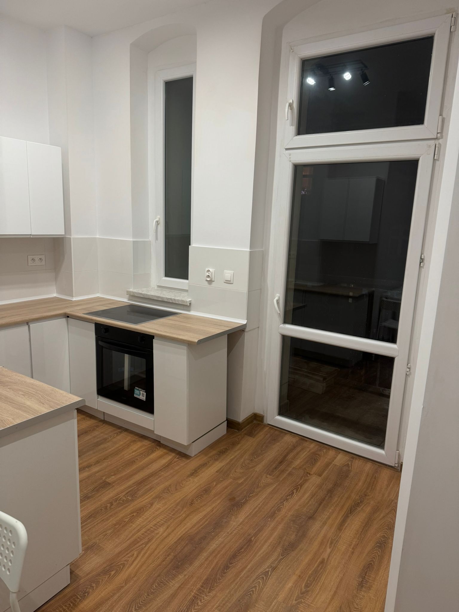 Nowoczesna kuchnia z białymi szafkami, drewnianymi blatami i podłogą z paneli imitujących drewno, widoczny fragment okna i drzwi balkonowych.