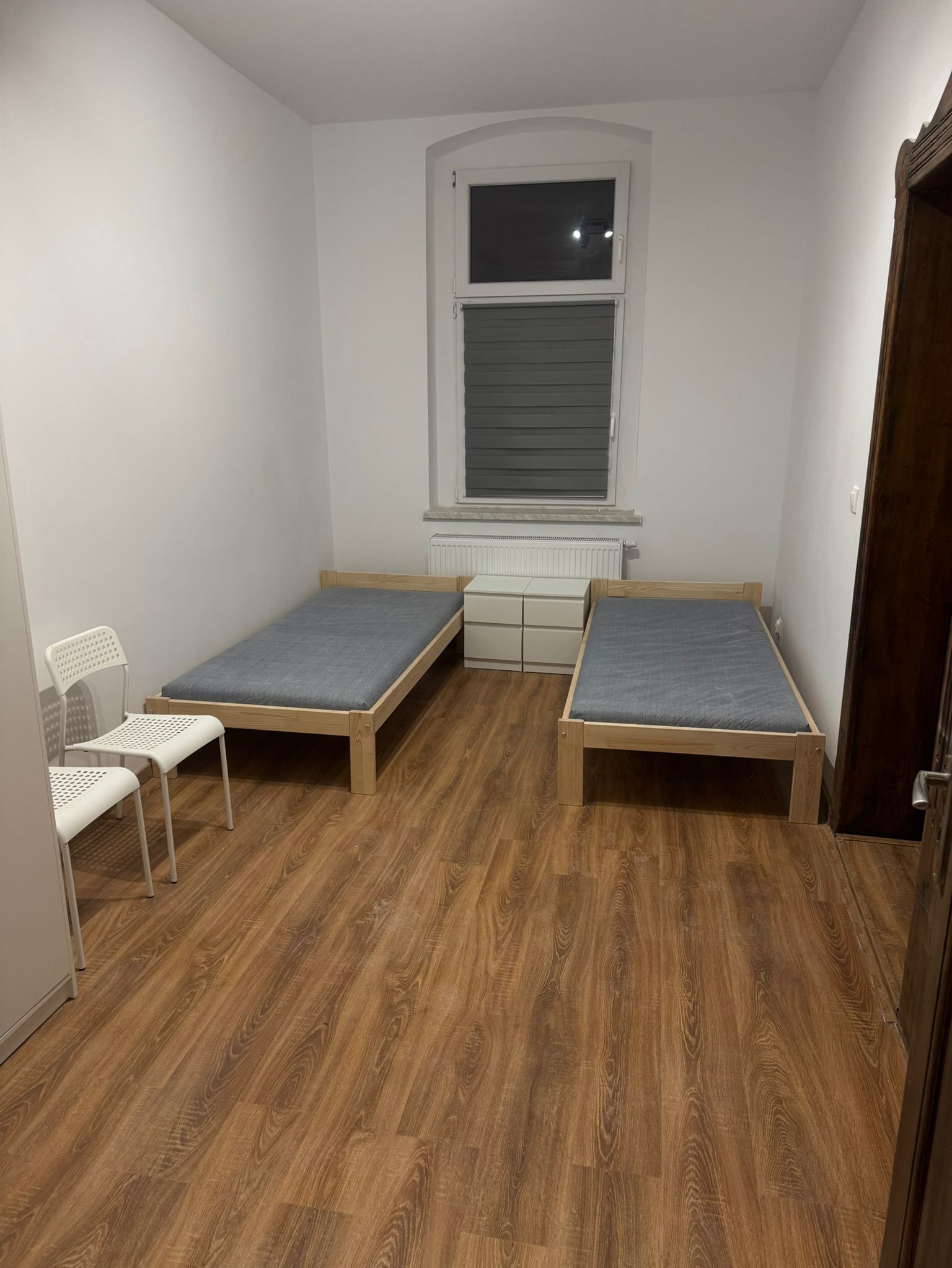 Pokój z dwoma drewnianymi łóżkami z szarymi materacami, białymi ścianami i panelami podłogowymi w odcieniach brązu, widok na białą komodę i okno z żaluzjami.