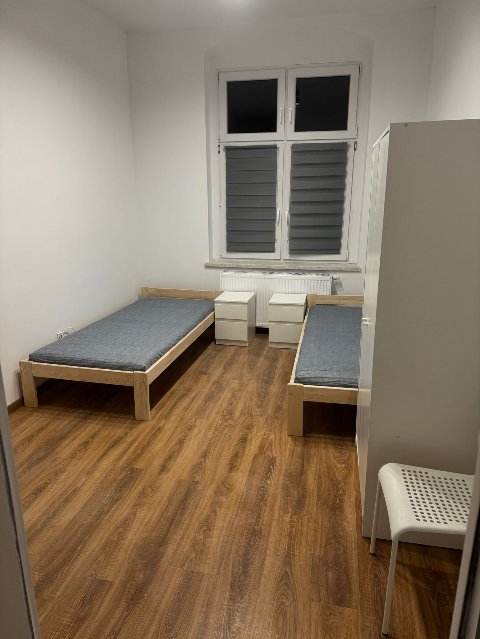 Pokój z dwoma drewnianymi łóżkami z szarymi materacami, dwiema białymi szafkami nocnymi, białą szafą i krzesłem na tle paneli podłogowych w odcieniach brązu.