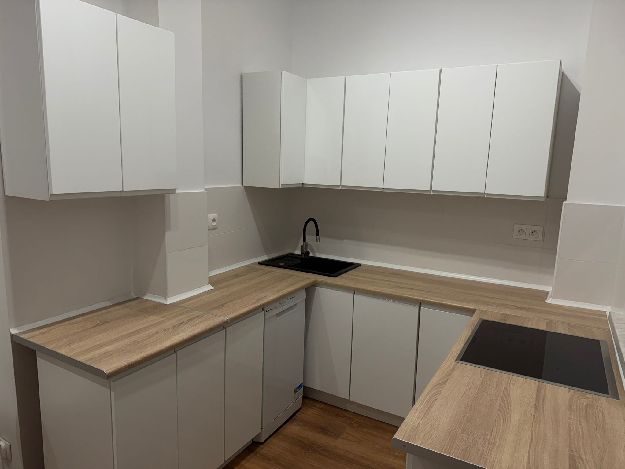 Nowoczesna kuchnia z białymi szafkami i blatem imitującym drewno, czarny zlew i płyta indukcyjna, widoczna zmywarka Candy.