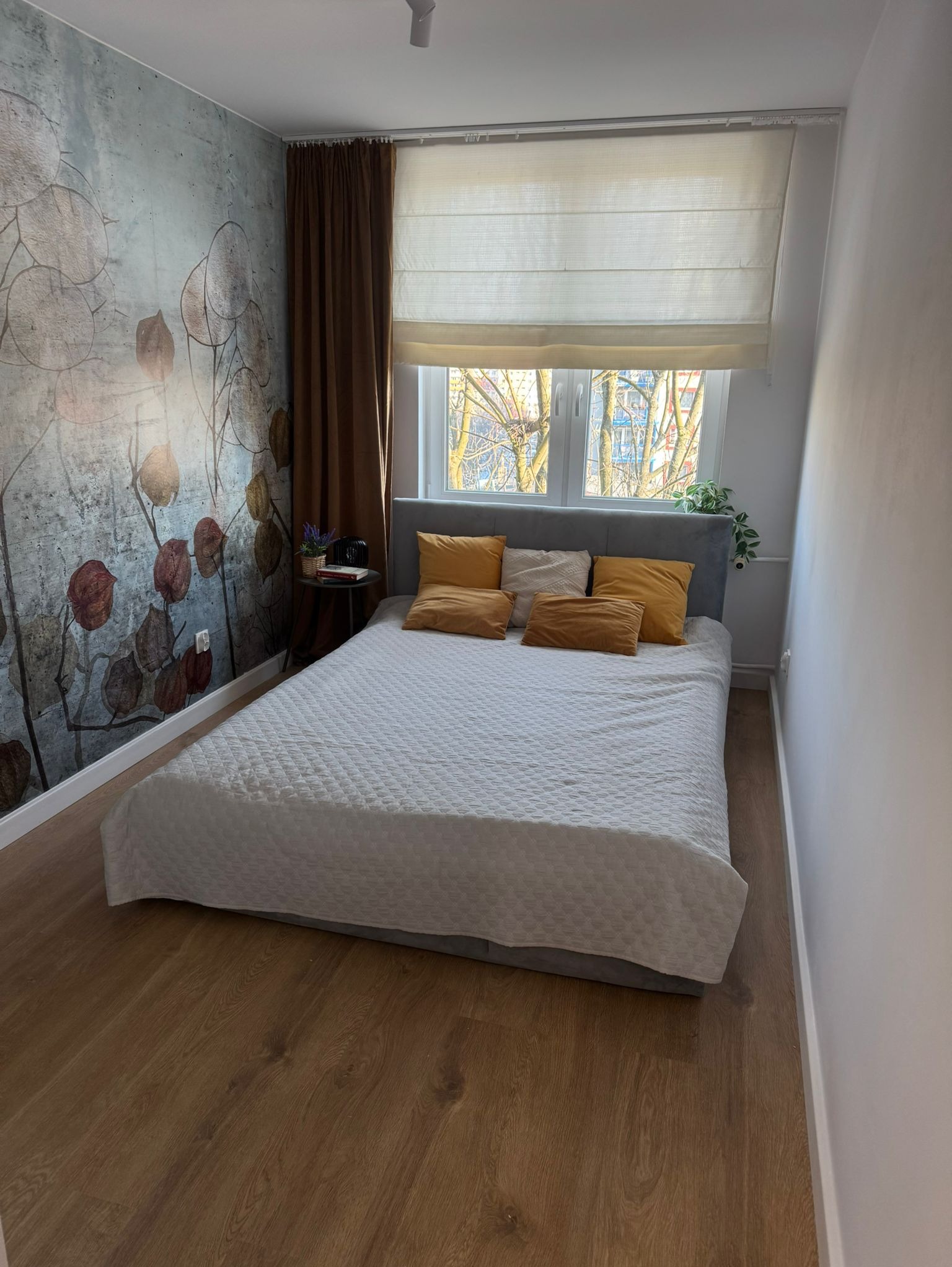 Sypialnia z drewnianą podłogą, łóżkiem z szarą ramą i białą narzutą, okno z roletą rzymską, ściana z dekoracyjnym wzorem roślinnym i brązowa zasłona.