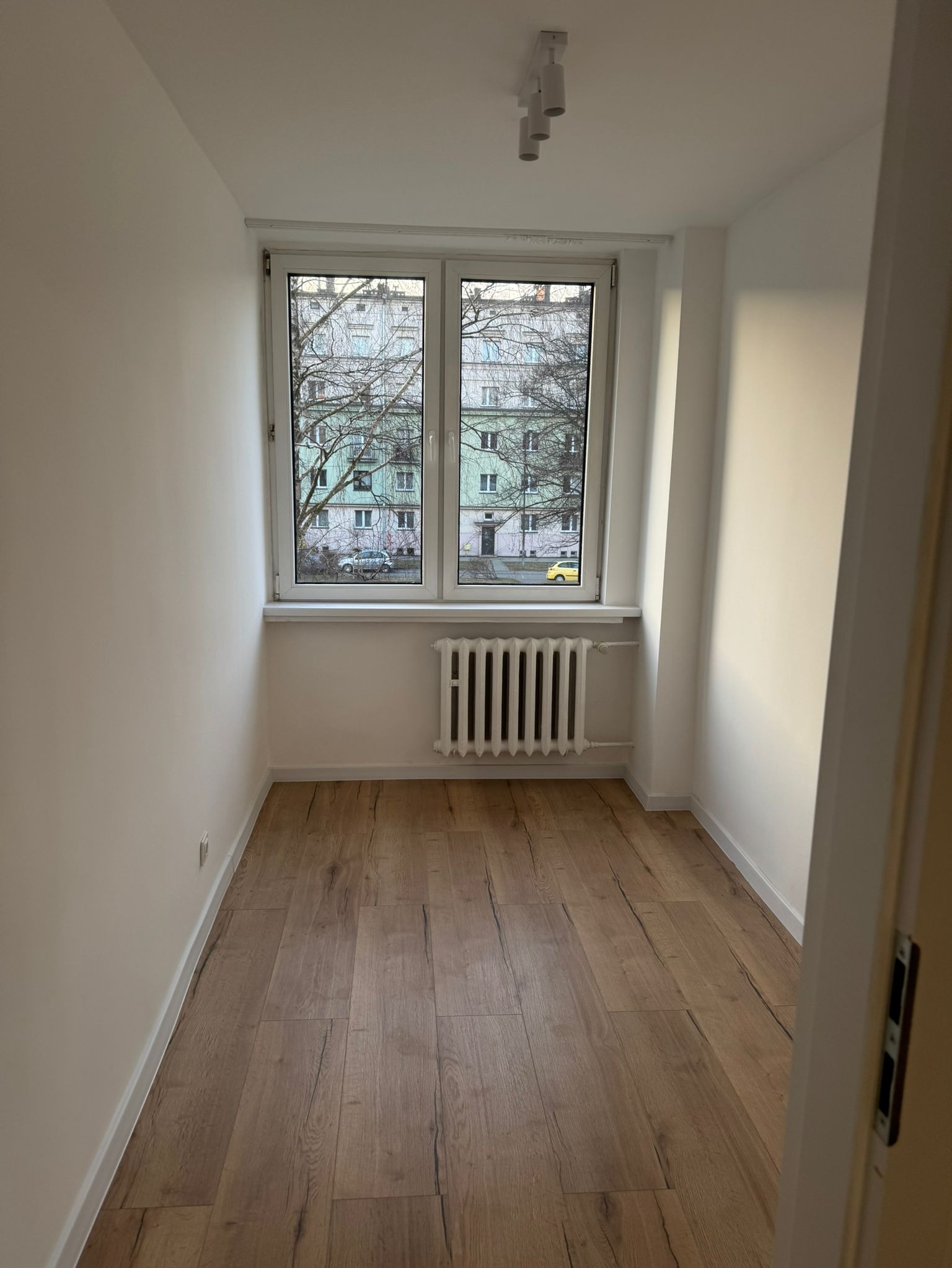 Pusty pokój po remoncie: panele podłogowe w odcieniach brązu, białe ściany, okno z widokiem na budynek i drzewa, grzejnik pod oknem, potrójna lampa sufitowa.