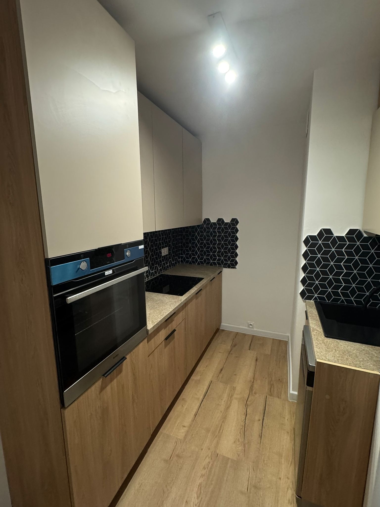 Wąska kuchnia z drewnopodobnymi panelami podłogowymi, szafkami z beżowymi i drewnianymi frontami oraz czarnymi heksagonalnymi płytkami nad blatem i przy zlewie.