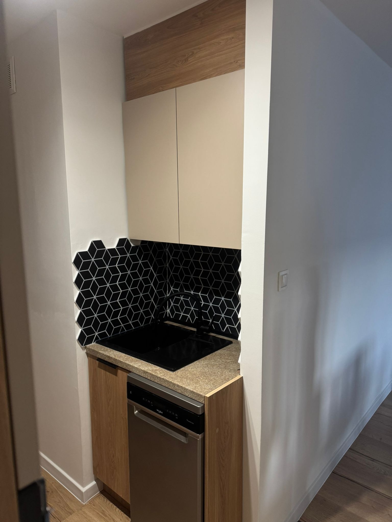 Nowoczesna kuchnia z czarnymi heksagonalnymi płytkami na ścianie, jasnymi szafkami i drewnianym blatem. Uwagę przykuwają geometryczne wzory płytek kontrastujące z gładkimi powierzchniami.