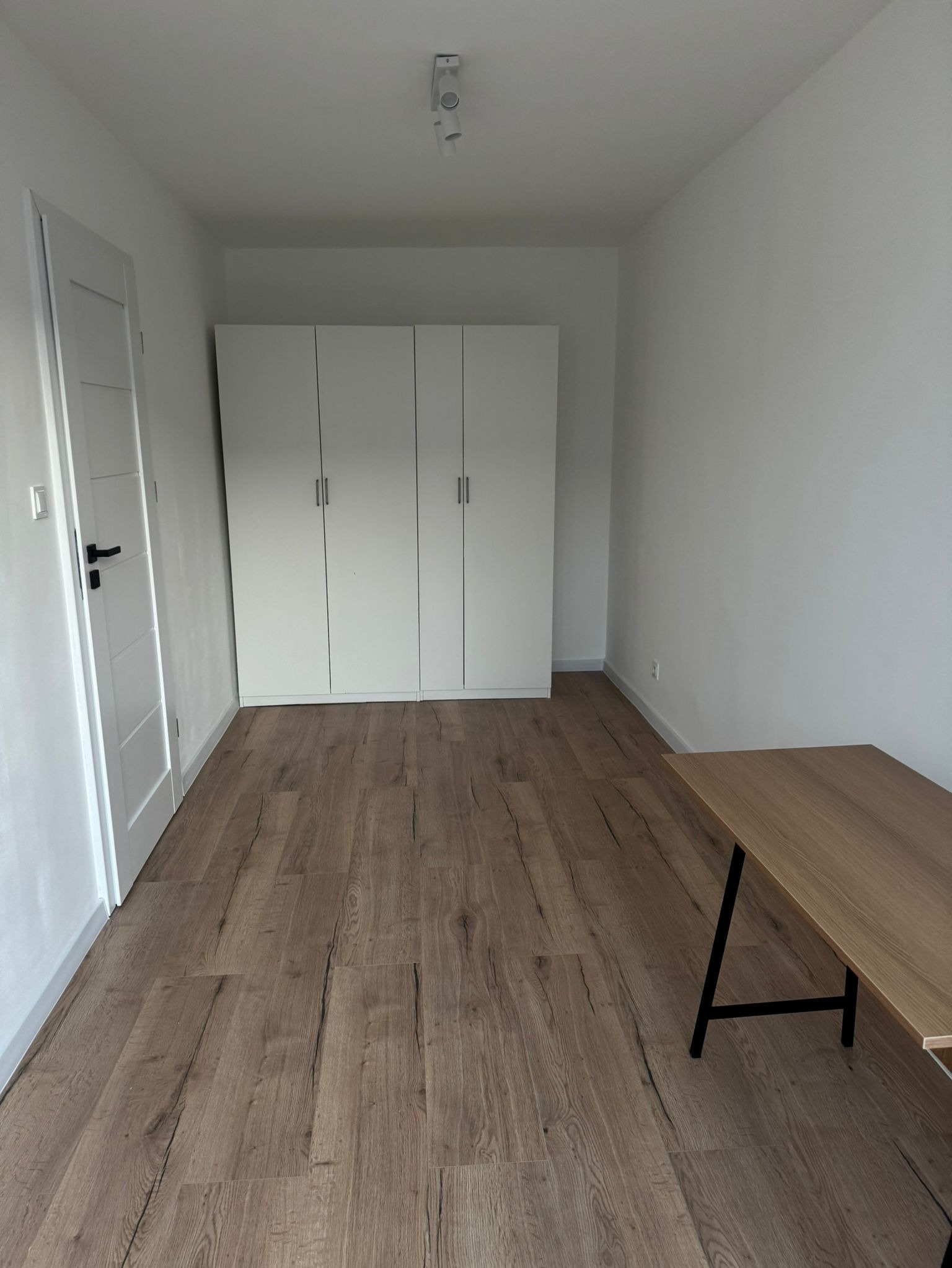 Pokój z ułożonymi panelami podłogowymi w odcieniach brązu, białymi ścianami, drzwiami z czarną klamką i czterodrzwiową szafą oraz biurkiem z drewnianym blatem na metalowych nogach.