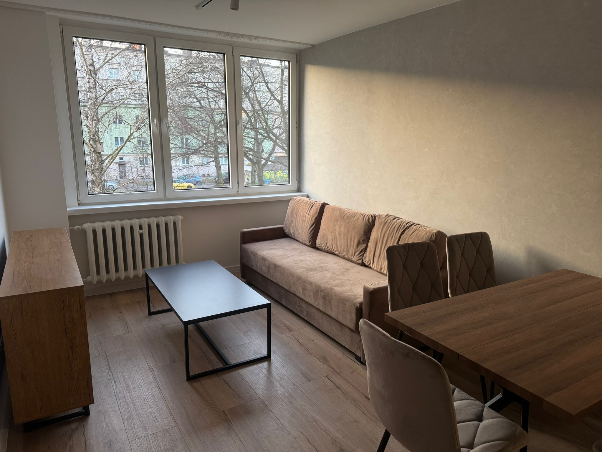 Jasne wnętrze pokoju dziennego z drewnianą podłogą, beżową sofą, stolikiem kawowym na czarnych nogach i stołem jadalnym z krzesłami, widok na drzewa za oknem.