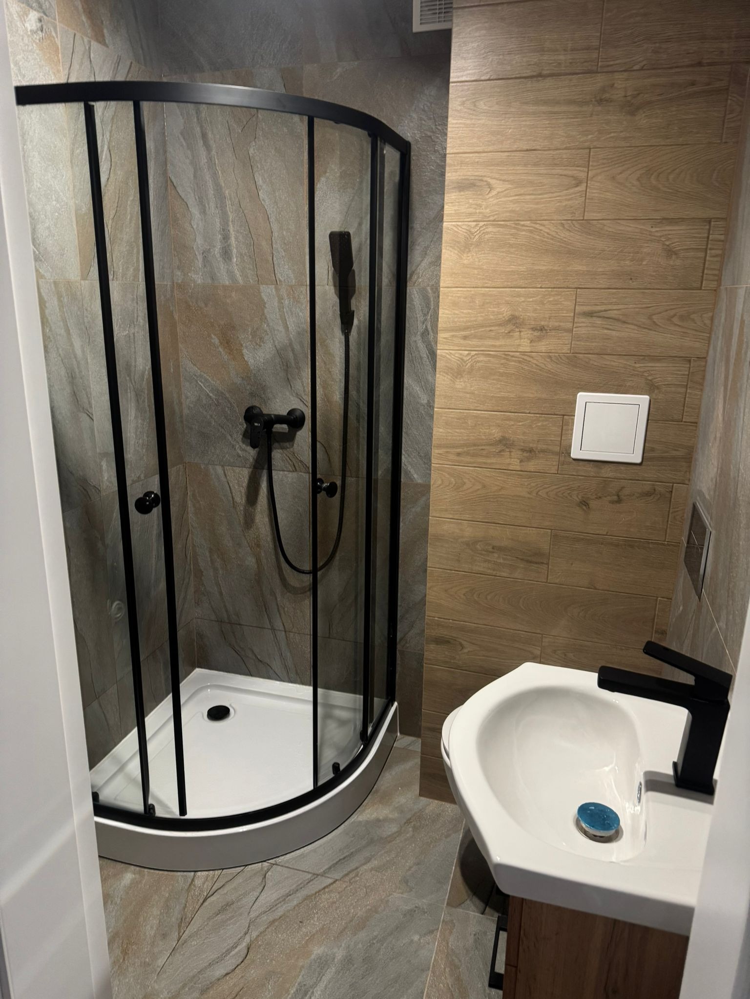 Nowoczesna łazienka z kabiną prysznicową z czarnymi ramami, umywalką z czarną armaturą i płytkami imitującymi drewno na ścianie.