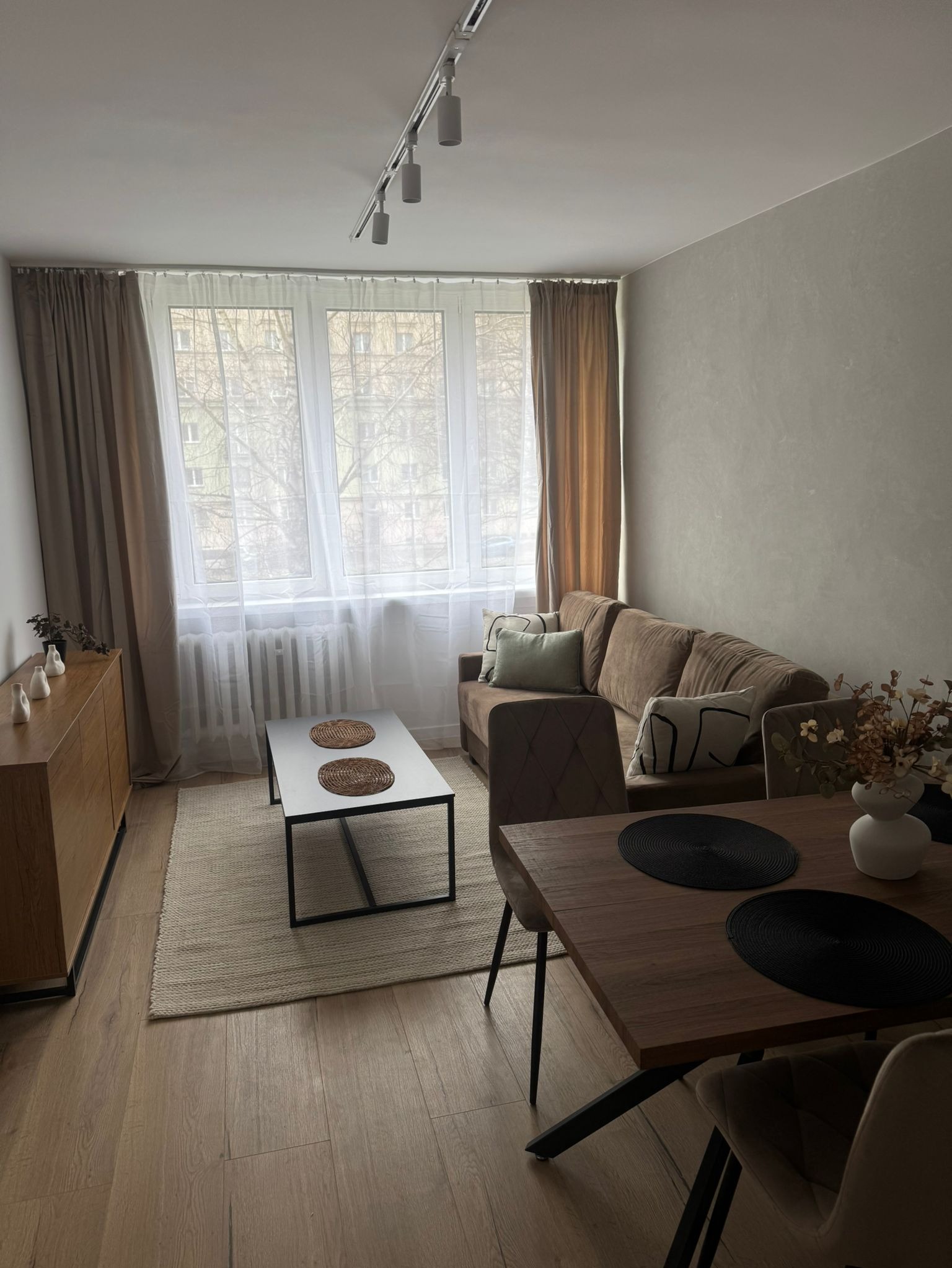 Jasne wnętrze salonu z drewnianą podłogą, brązową sofą, stolikiem kawowym z rattanowymi podkładkami, drewnianym stołem jadalnym z czarnymi podkładkami, oświetleniem sufitowym na szynie i zasłonami...