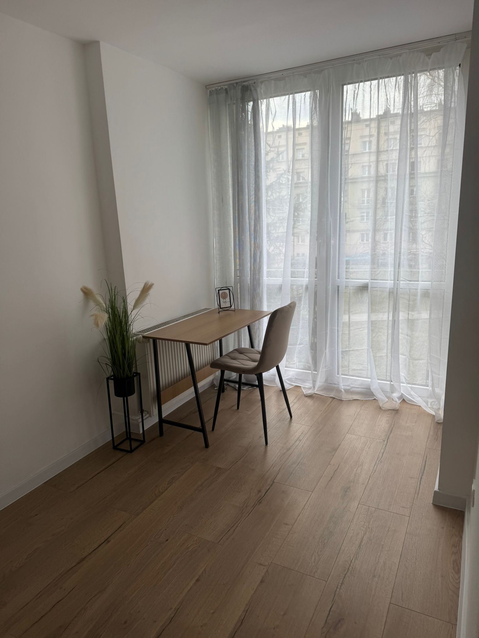 Jasne wnętrze pokoju z drewnianą podłogą, biurkiem przy oknie zasłoniętym białymi, zwiewnymi firankami i krzesłem obitym beżową tkaniną. Na podłodze widoczne panele podłogowe ułożone w jodełkę.