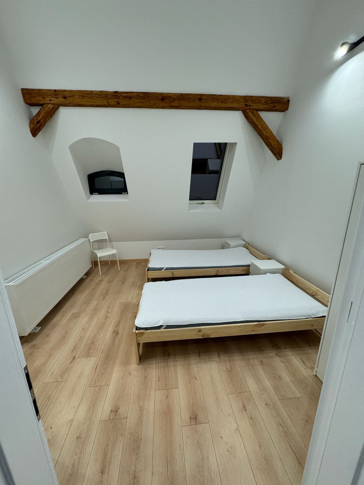 Poddasze z dwoma drewnianymi łóżkami z białymi materacami, drewnianą podłogą imitującą panele, białym grzejnikiem, krzesłem i widocznymi belkami stropowymi.