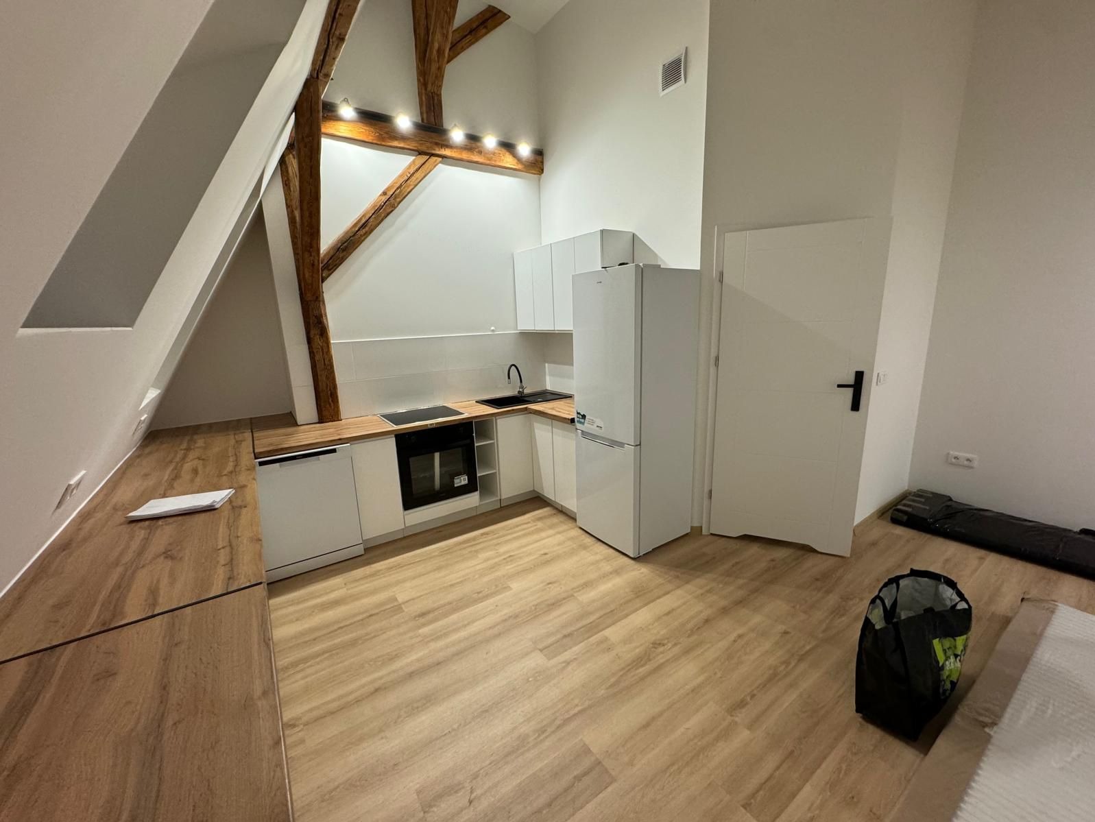 Nowoczesna kuchnia w odnowionym wnętrzu na poddaszu z drewnianymi belkami stropowymi, białymi meblami i panelami podłogowymi w odcieniach drewna.