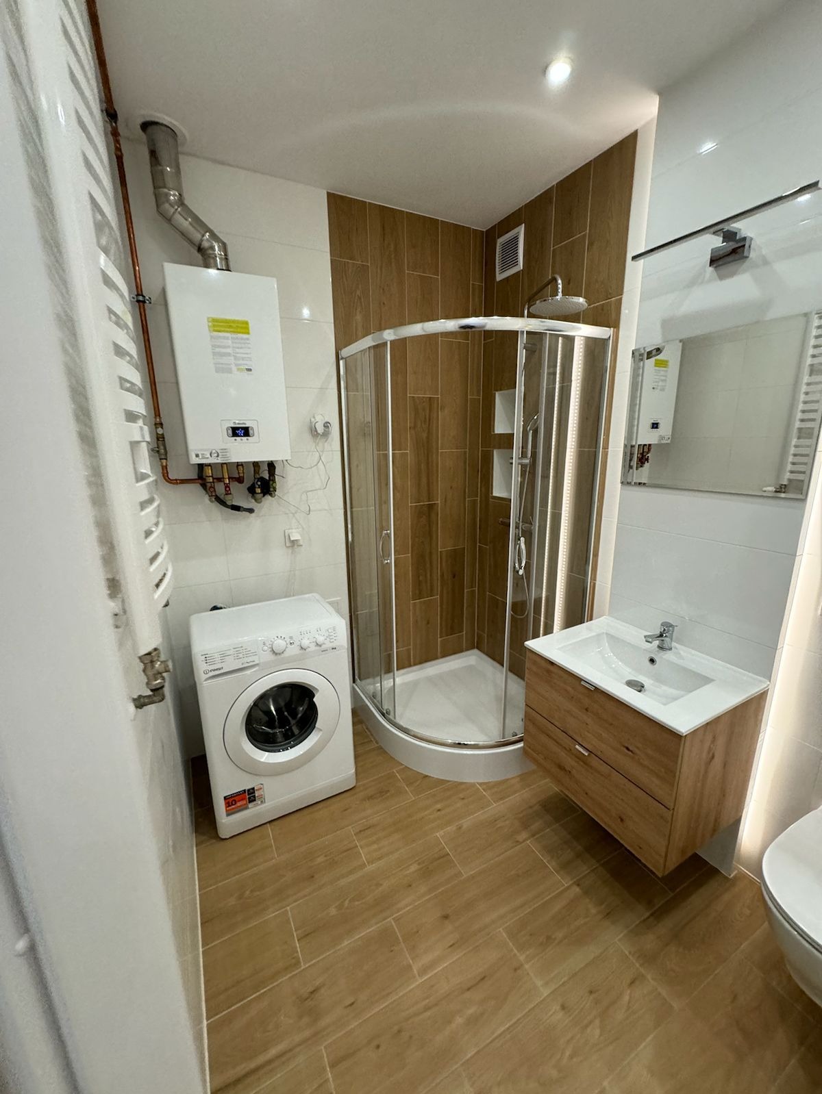 Ukończona łazienka z drewnopodobnymi płytkami, kabiną prysznicową, umywalką z szafką w stylu skandynawskim, pralką i piecem gazowym z odprowadzeniem spalin.
