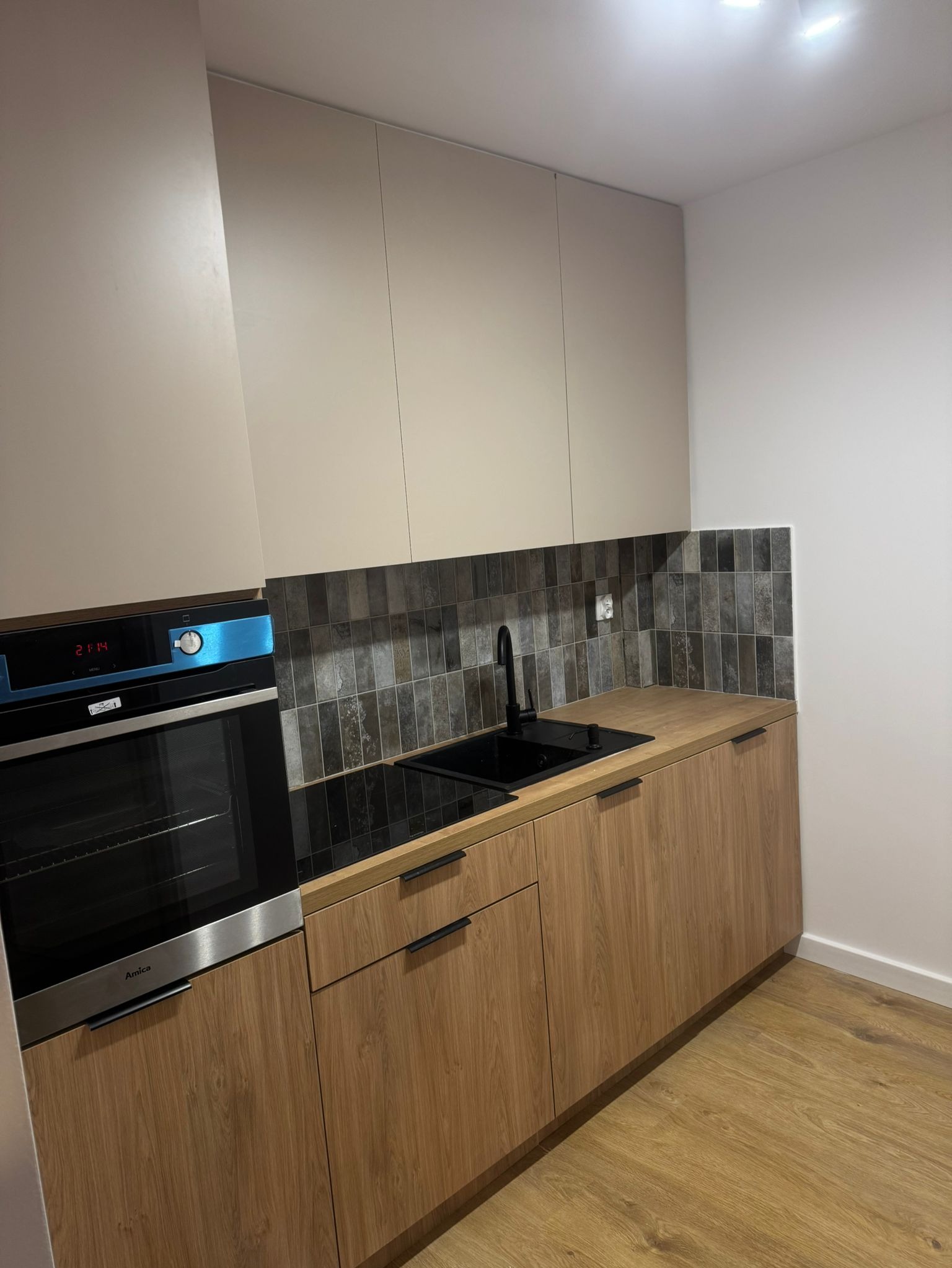 Nowoczesna kuchnia z drewnianymi szafkami dolnymi i beżowymi górnymi, czarnym zlewem i baterią, płytkami imitującymi kamień na ścianie oraz piekarnikiem marki Amica.