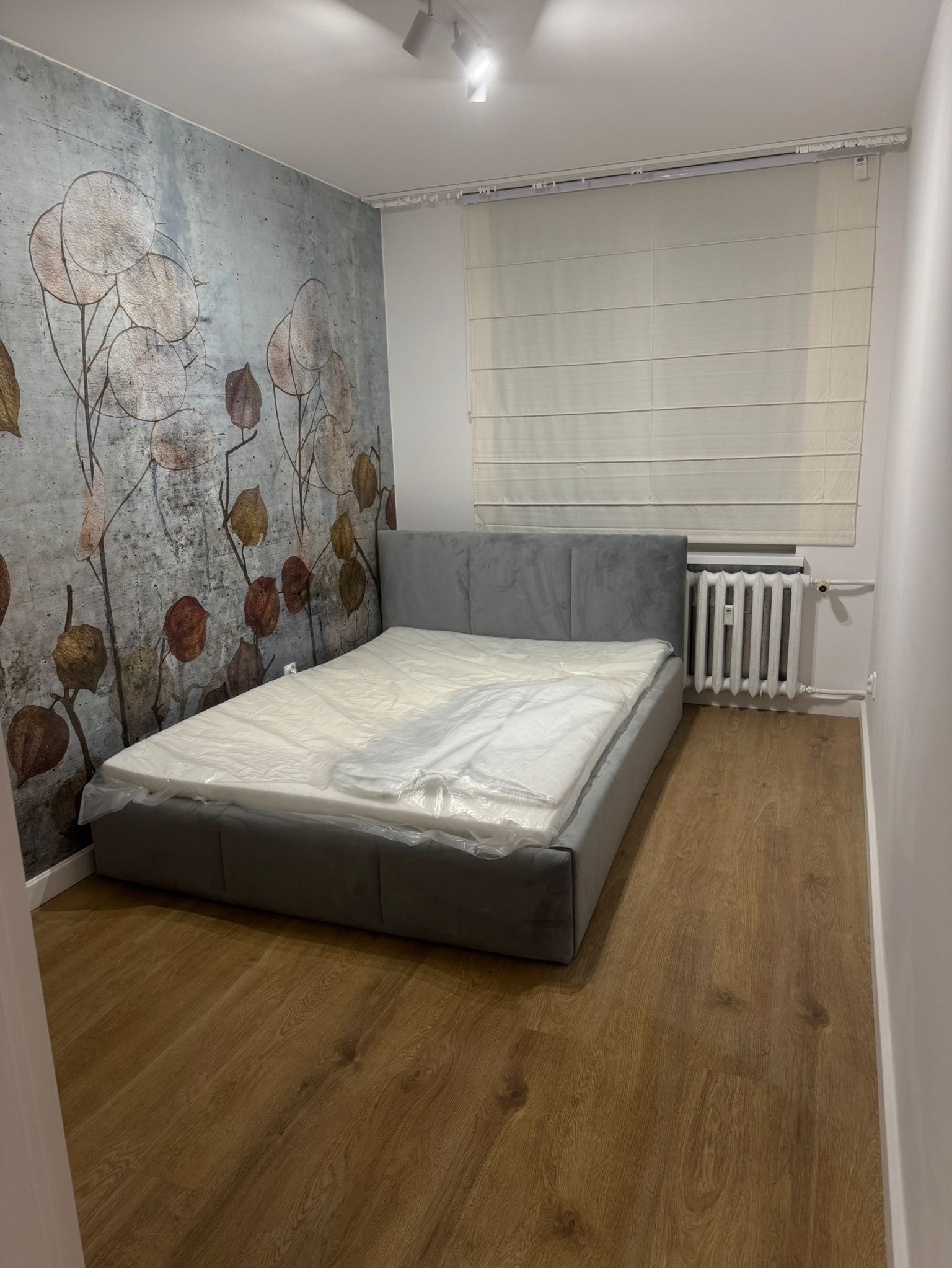 Nowe panele podłogowe w sypialni z szarym łóżkiem, ściana z motywem roślinnym, grzejnik pod oknem z roletami rzymskimi.