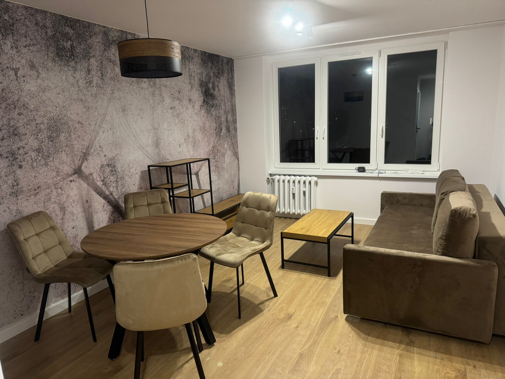 Jasne wnętrze salonu z okrągłym stołem, krzesłami z tapicerką w odcieniach beżu, brązową sofą i lampą wiszącą z drewnianym akcentem, na podłodze panele podłogowe o jasnym odcieniu drewna, ściana...