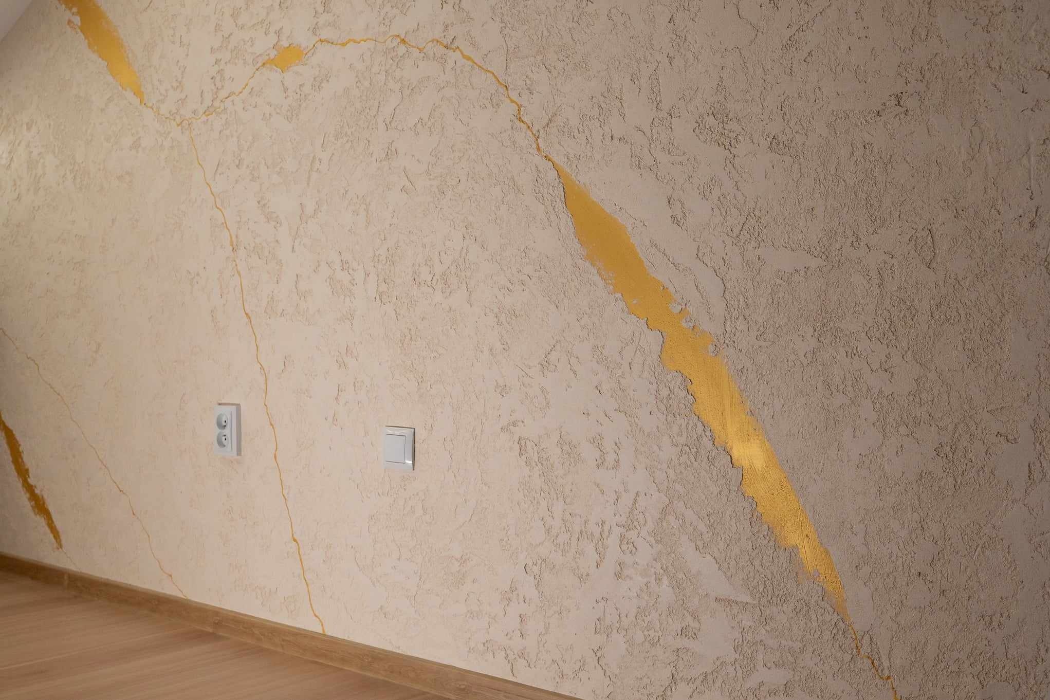 Dekoracyjna ściana z beżową, chropowatą fakturą i złotymi, nieregularnymi żyłkami, widoczne gniazdka elektryczne.