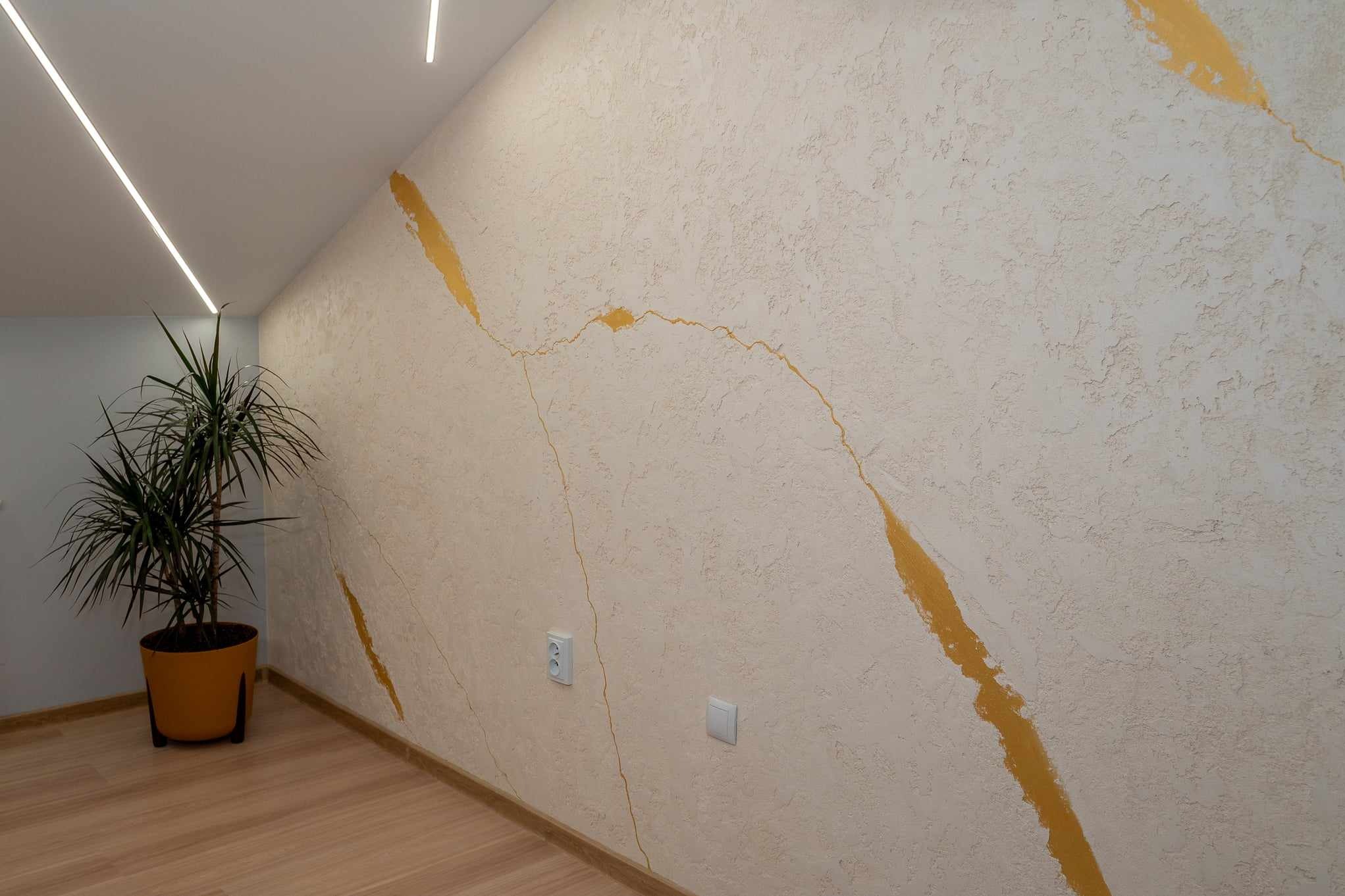Teksturowana ściana w odcieniach beżu z nieregularnymi, złotymi akcentami, oświetlona listwą LED, z drewnianą podłogą i rośliną doniczkową w narożniku.