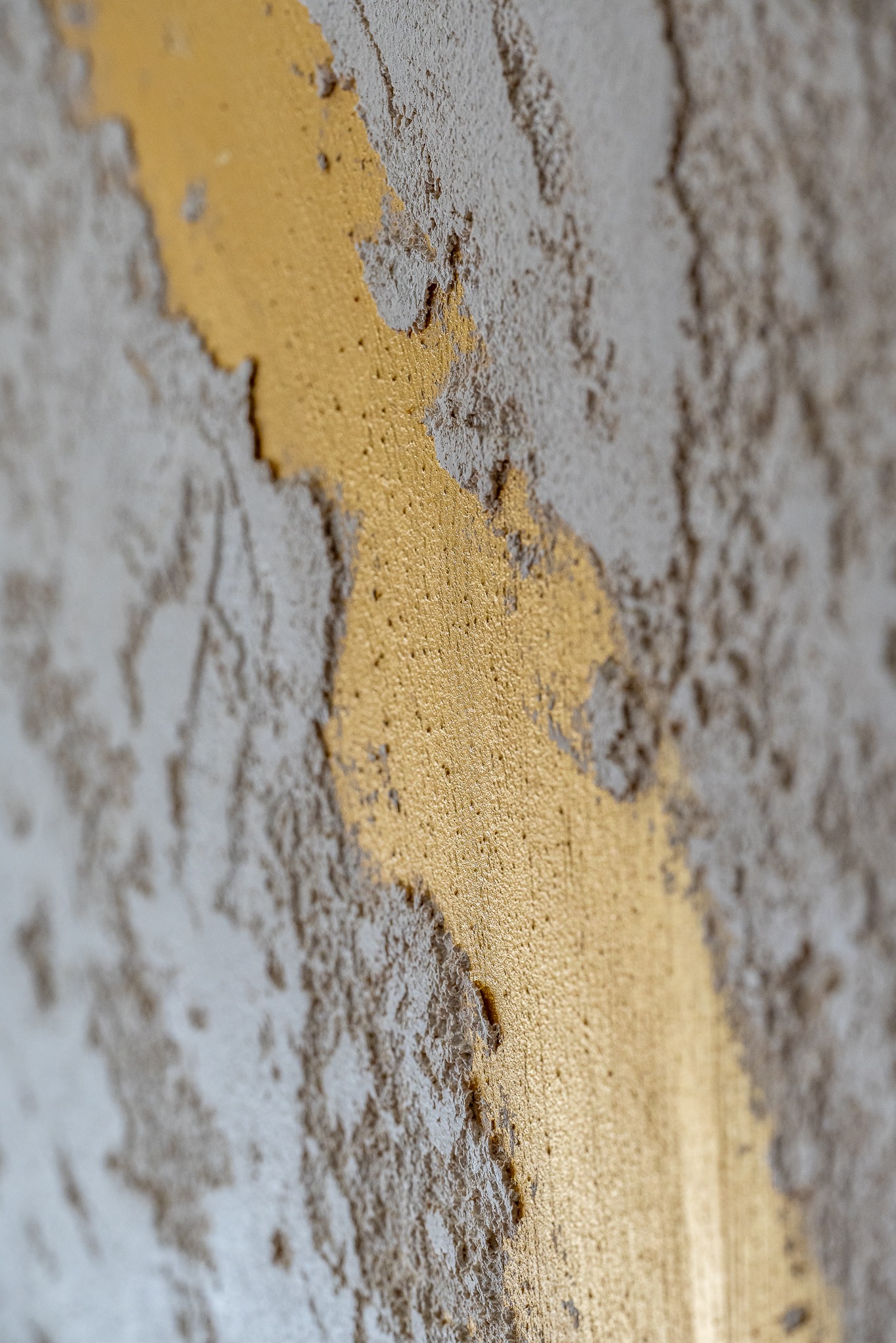 Dekoracyjna ściana z szarego szpachlowania z pionowym pasem złotej farby, widoczne nierówności i faktura.