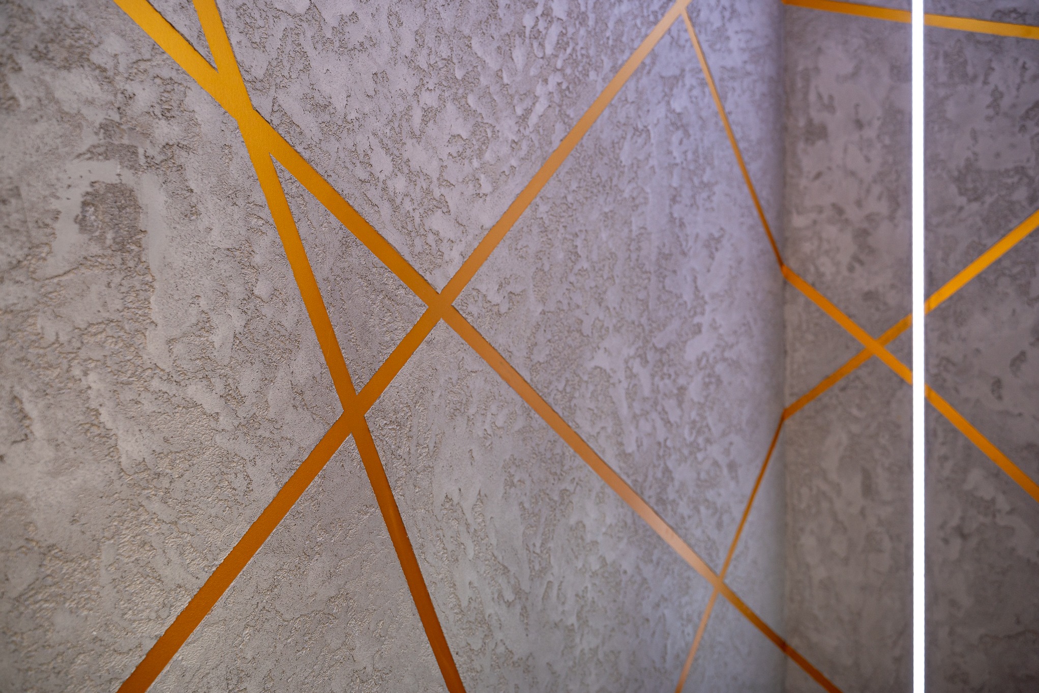 Dekoracyjna ściana z szarego tynku strukturalnego z geometrycznym wzorem z pomarańczowych pasków, oświetlona pionową lampą LED.