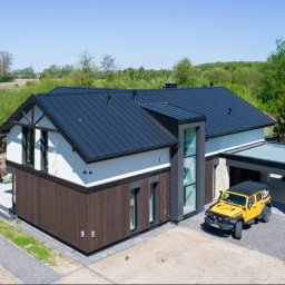 Kamil Melon MELKAM-BUD - Nowoczesny dom z ciemnym dachem i drewnianą elewacją, żółty samochód terenowy zaparkowany przy wiacie, widok z lotu ptaka.