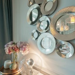 Kamil Melon MELKAM-BUD - Dekoracyjna ściana z zestawem okrągłych luster o srebrnych, młotkowanych ramach, odbijających wnętrze z designerskimi lampami, uzupełniona bukietem hortensji i nowoczesną szklaną lampą kulistą.