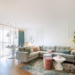 Kamil Melon MELKAM-BUD - Jasne wnętrze salonu z narożną sofą w odcieniach szarości, okrągłym stołem z przezroczystymi krzesłami, dywanem w pastelowych kolorach i dekoracyjnymi elementami na ścianie.