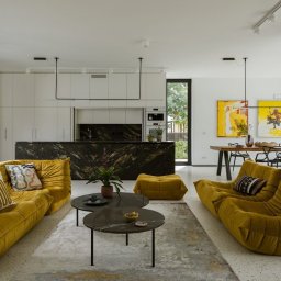 Kamil Melon MELKAM-BUD - Przestronny salon z żółtymi sofami modułowymi, marmurowym blatem kuchennym i minimalistycznym oświetleniem, dopełniony abstrakcyjnymi obrazami i roślinami doniczkowymi.