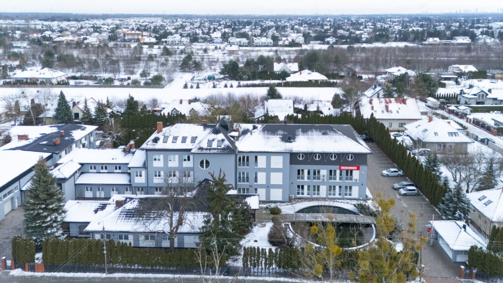 Widok z góry na szary budynek z napisem 'minimax' na elewacji w zimowej scenerii, otoczony śniegiem pokrytymi dachami domów i drzewami.