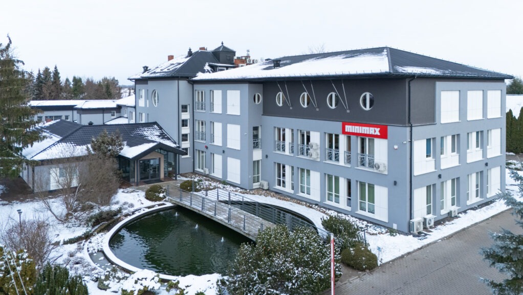 Nowoczesny, trzypiętrowy budynek biurowy w szaro-białej kolorystyce z logo Minimax, otoczony zimowym krajobrazem z oczkiem wodnym i mostkiem.