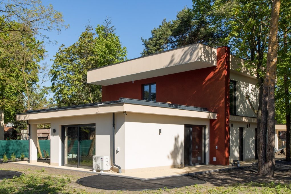 Nowoczesny dwukondygnacyjny dom z płaskim dachem, elewacją w kolorze białym i ceglastej czerwieni, otoczony zielenią drzew i częściowo przygotowanym terenem.