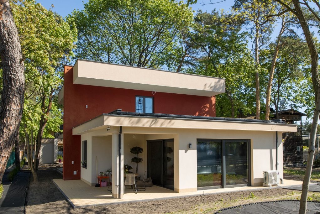 Nowoczesny dom z płaskim dachem, elewacją w kolorze terakoty i beżowym wykończeniem, otoczony zielenią drzew, widok z zewnątrz w słoneczny dzień.