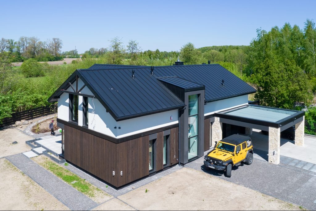 Nowoczesny dom z ciemnym dachem i drewnianą elewacją, żółty samochód terenowy zaparkowany przy wiacie, widok z lotu ptaka.