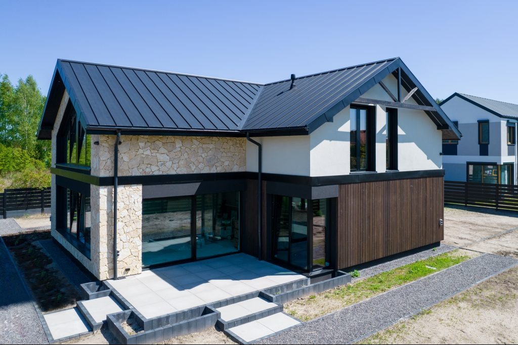 Nowoczesny dom jednorodzinny z elewacją z kamienia naturalnego, ciemną blachą na dachu i drewnianą okładziną, widok z góry na taras z płyt betonowych i ścieżkę z grysu.