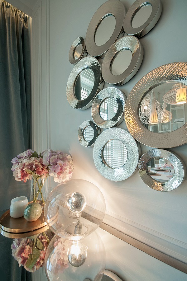 Dekoracyjna ściana z zestawem okrągłych luster o srebrnych, młotkowanych ramach, odbijających wnętrze z designerskimi lampami, uzupełniona bukietem hortensji i nowoczesną szklaną lampą kulistą.