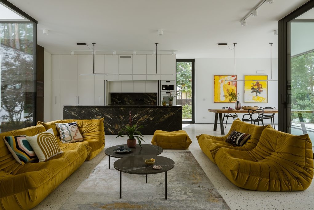 Przestronny salon z żółtymi sofami modułowymi, marmurowym blatem kuchennym i minimalistycznym oświetleniem, dopełniony abstrakcyjnymi obrazami i roślinami doniczkowymi.