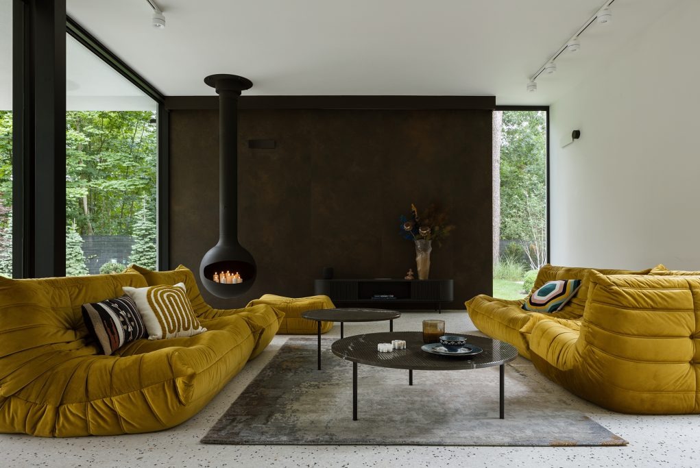 Nowoczesny salon z wiszącym kominkiem w kształcie kuli, żółtymi sofami modułowymi, ciemną ścianą akcentową oraz widokiem na zieleń za oknem.