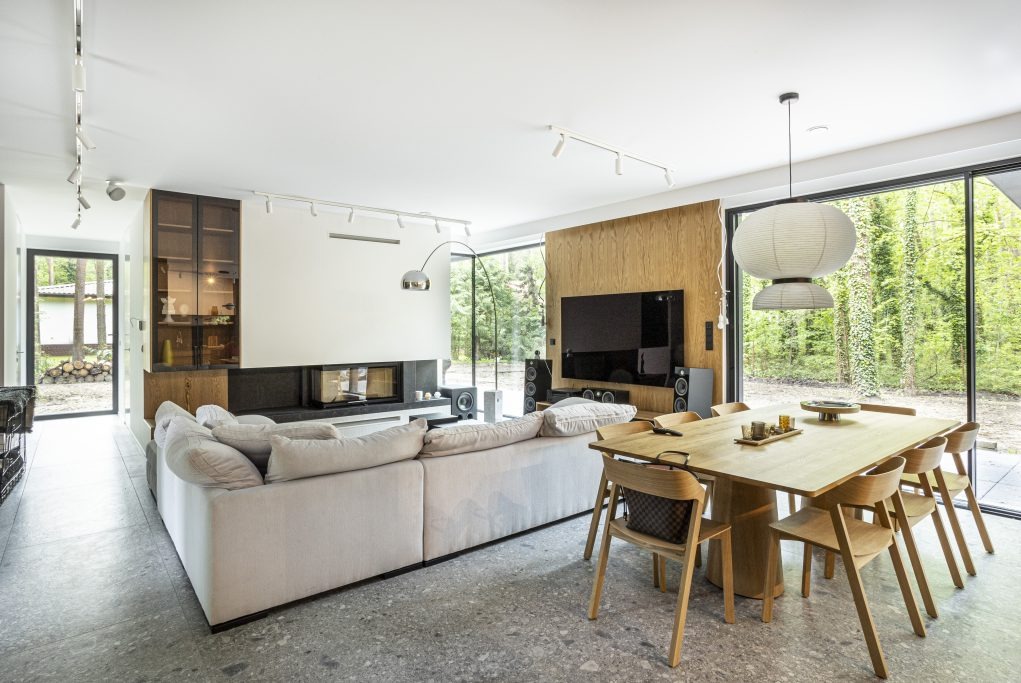 Jasny salon z dużym oknem na las, szarym lastryko na podłodze, beżową sofą narożną, drewnianym stołem jadalnym i kominkiem z czarnym obramowaniem.