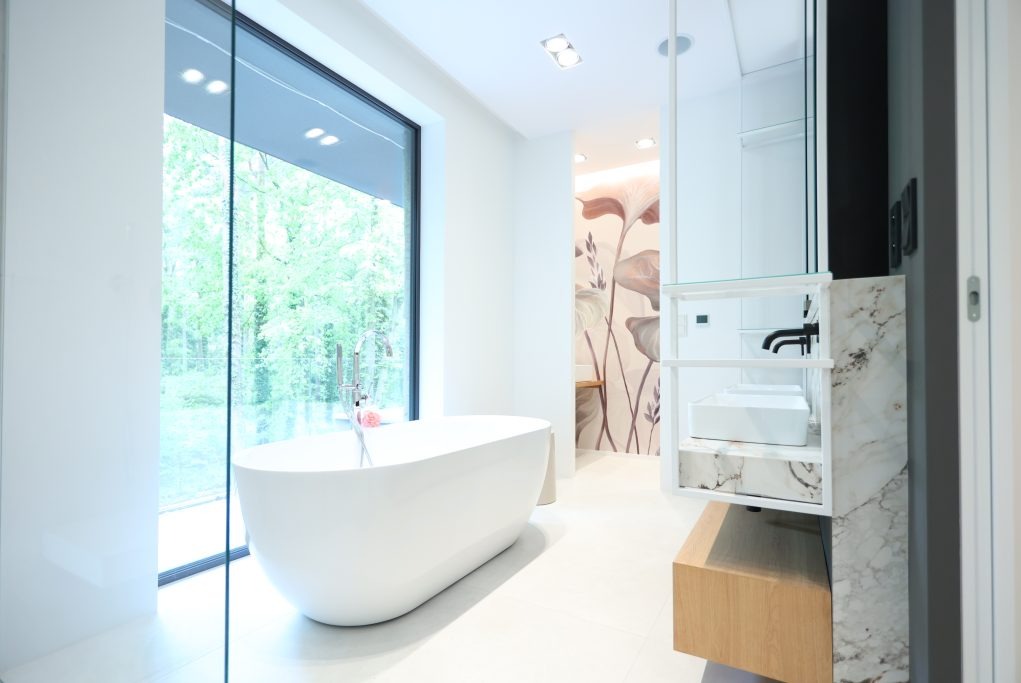 Nowoczesna, jasna łazienka z wolnostojącą wanną przy dużym oknie z widokiem na zieleń, minimalistyczną umywalką z marmurowym blatem i tapetą w motywy roślinne w tle.
