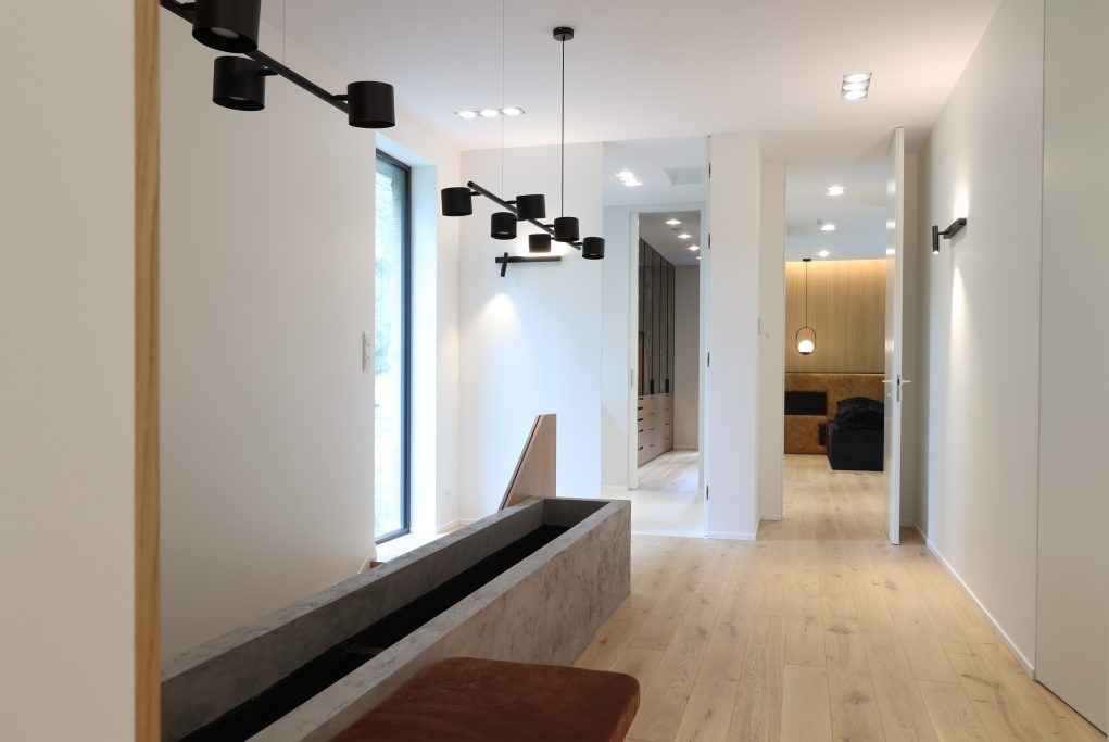 Nowoczesny przedpokój z drewnianą podłogą, betonową donicą i designerskimi lampami wiszącymi w kolorze czarnym, widok na garderobę i sypialnię w głębi.