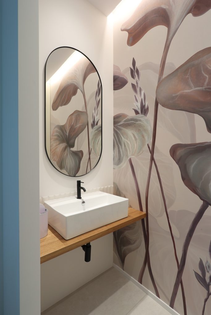 Nowoczesna łazienka z prostokątną umywalką nablatową, czarną armaturą i lustrem w czarnej ramie na tle ściany z motywem roślinnym w odcieniach beżu i brązu.