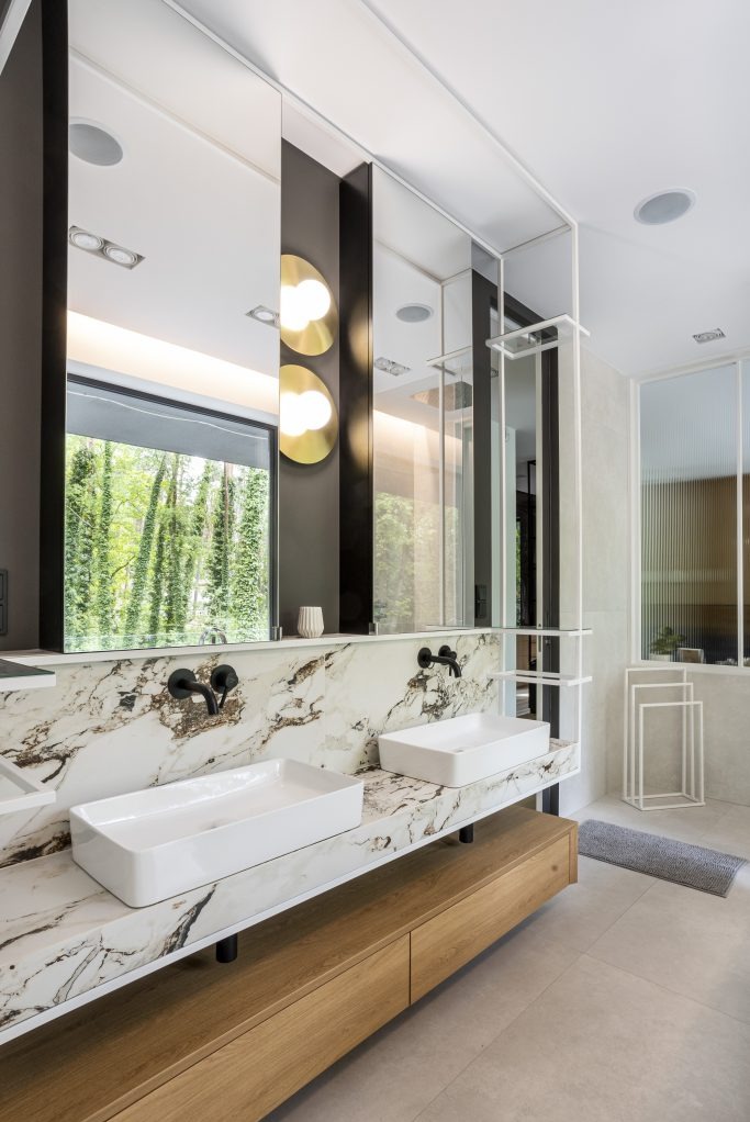 Nowoczesna łazienka z dwoma prostokątnymi umywalkami nablatowymi na blacie z marmuru o wyrazistym wzorze, czarne baterie, lustra z podświetleniem i widokiem na zieleń za oknem, drewniana szafka...