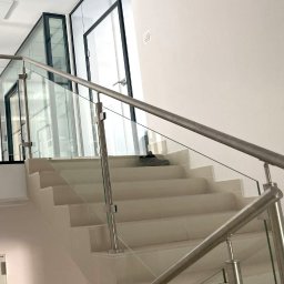 Loftowe-Design - Schody wyłożone jasnymi płytkami z balustradą ze stali nierdzewnej i szkła, widoczne buty na schodach, w tle przeszklone drzwi z czarnymi ramami.