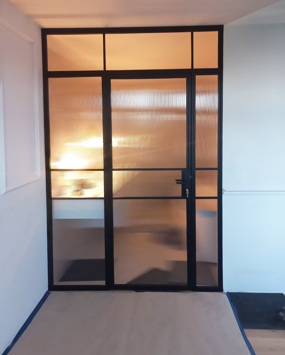 Szklane drzwi z czarną ramą i ryflowanym szkłem, widoczne w jasnym wnętrzu z białymi ścianami i zabezpieczoną podłogą.