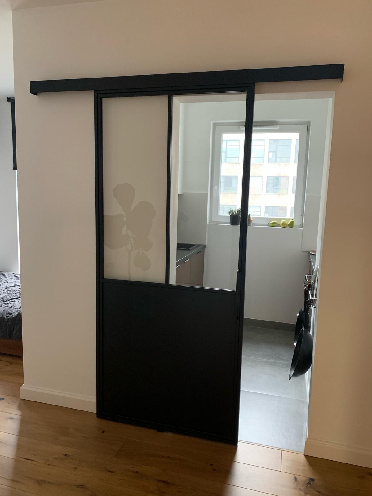 Czarne, przesuwne drzwi z częściowo matowym szkłem z motywem roślinnym, oddzielające pokój od kuchni w mieszkaniu, widoczne drewniane podłogi i jasne ściany.