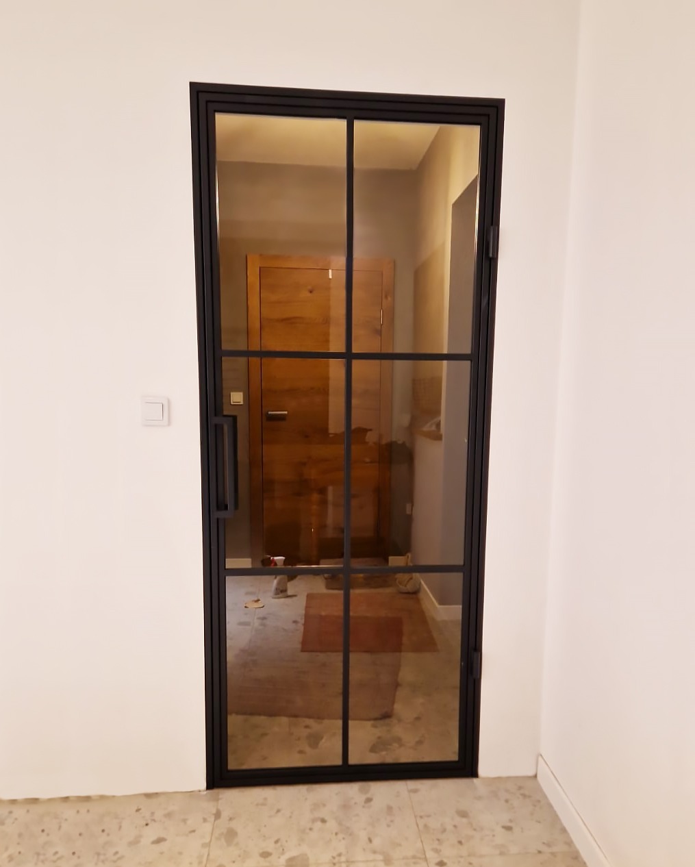 Nowoczesne drzwi z czarną ramą i szklanymi panelami, widok na wnętrze z drewnianymi drzwiami w tle.