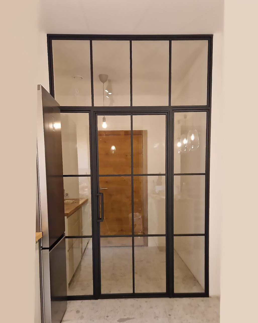 Przeszklona konstrukcja z czarnymi szprosami oddzielająca pomieszczenia, widoczna kuchnia i drewniane drzwi w tle.