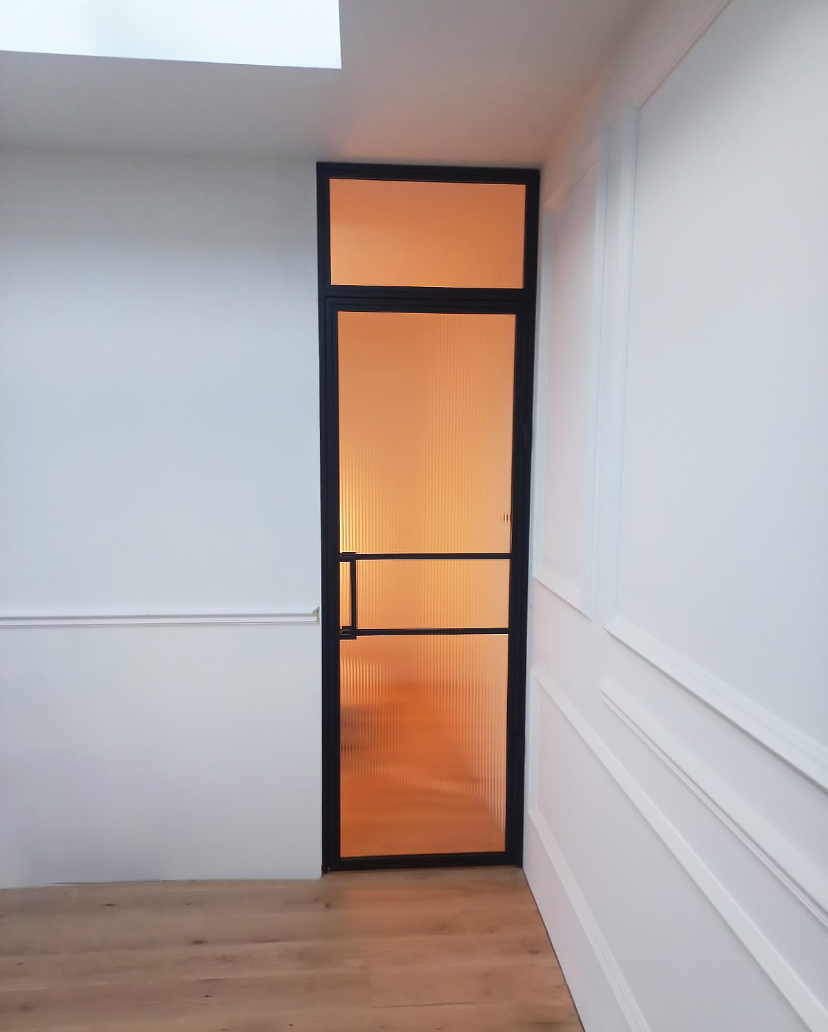 Wysoka, wąska ścianka działowa z żebrowanego szkła w czarnej ramie, oddzielająca przestrzeń, widok na fragment podłogi i sufit z oknem dachowym.