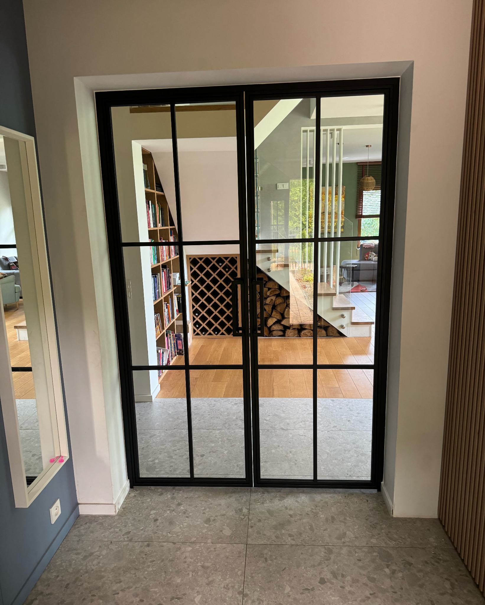 Dwuskrzydłowe drzwi z czarnymi szprosami oddzielają korytarz od salonu z biblioteczką i schodami z drewnem pod stopniami.