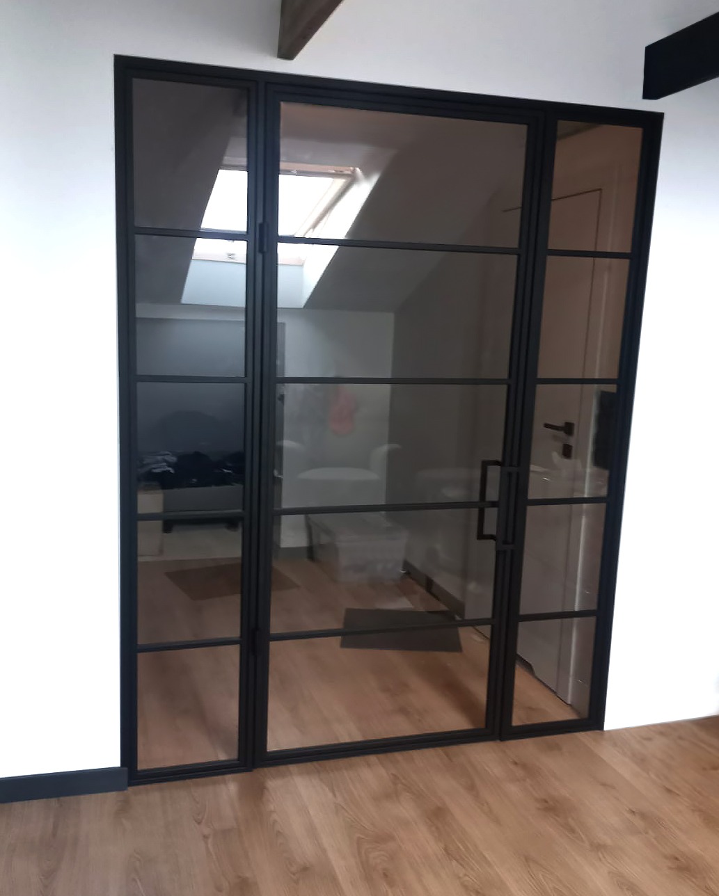 Nowoczesne, dwuskrzydłowe drzwi z czarnymi szprosami i przezroczystym szkłem, dzielące przestrzeń poddasza z oknem dachowym i pomieszczenia z drzwiami.