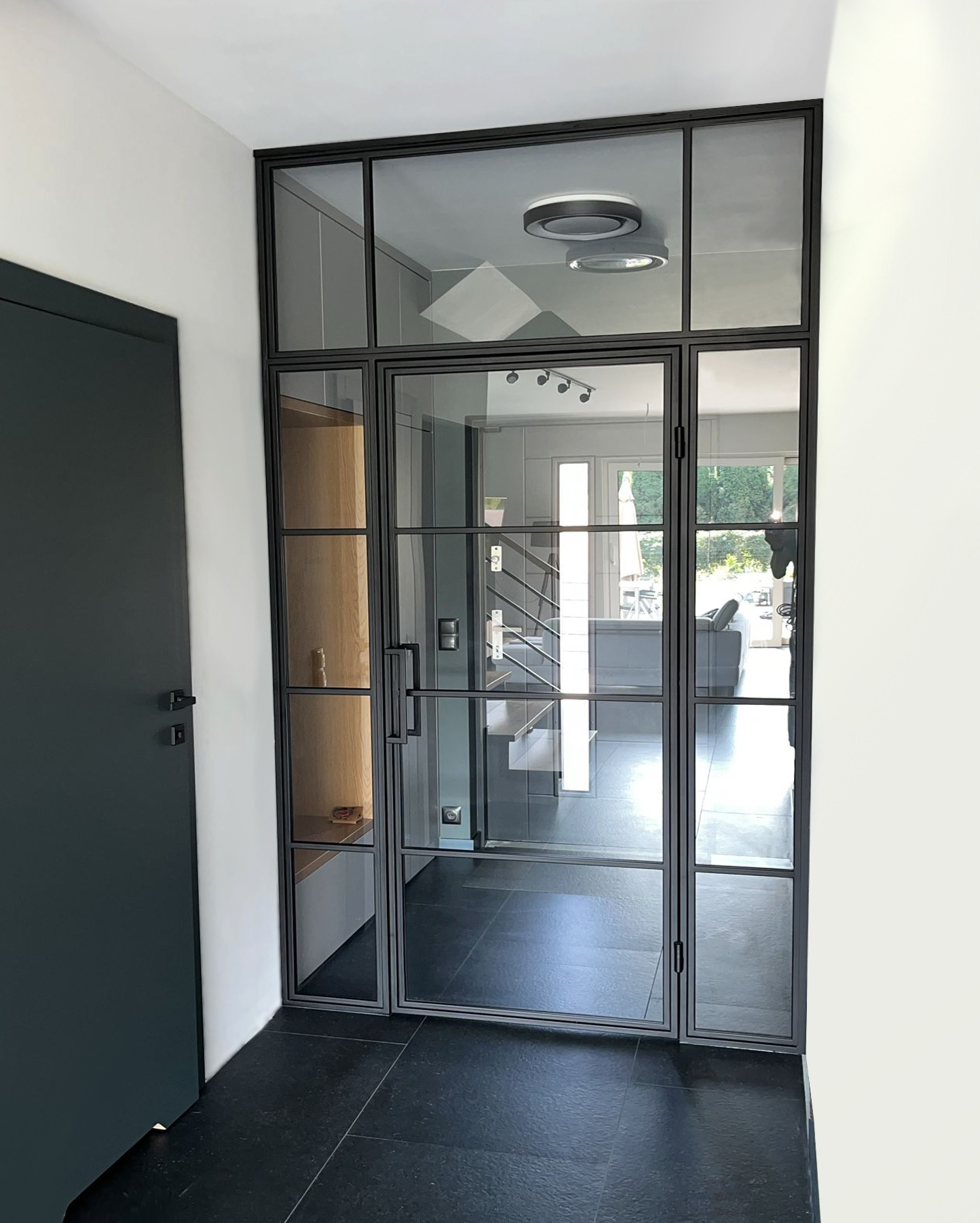 Szklane drzwi wewnętrzne z czarną ramą w stylu loft, widok na schody i salon, czarna podłoga z płytek.